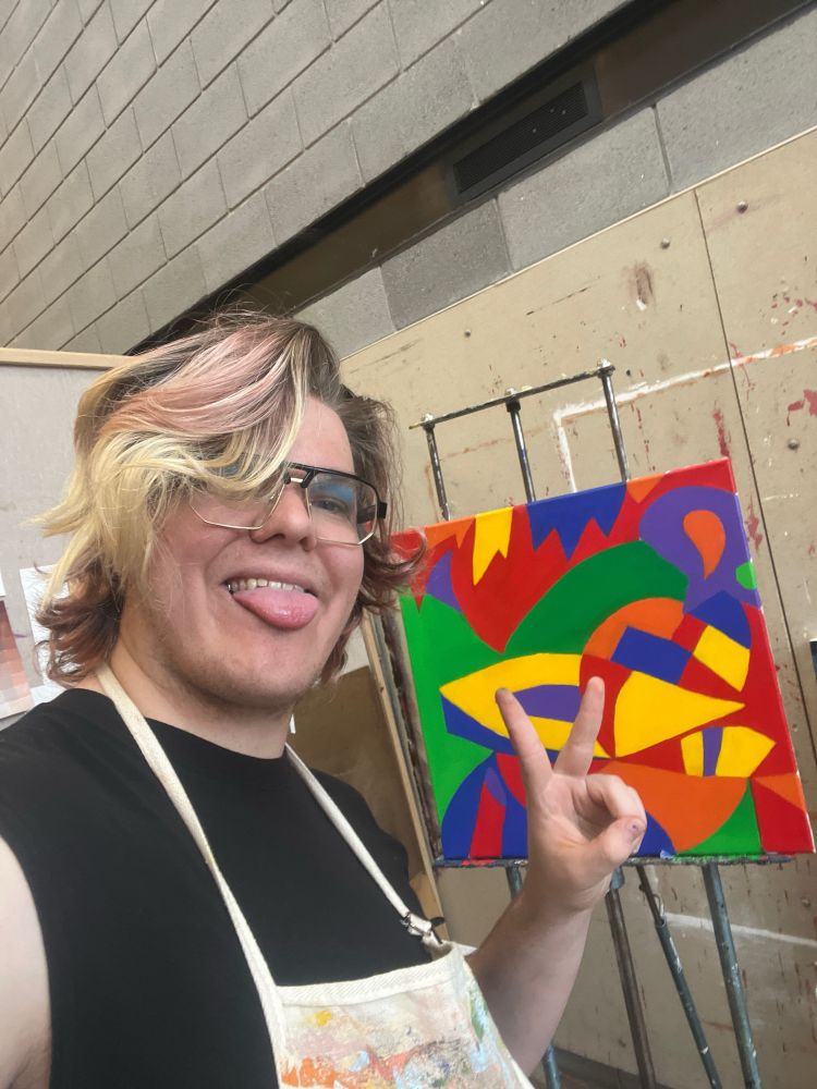 An artist posing in font of a painting in their studio, smiling and making a peace sign.