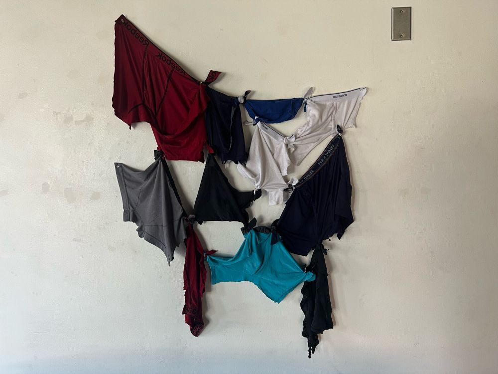 An assemblage of mens underwear scraps knotted together and pinned to a white wall.