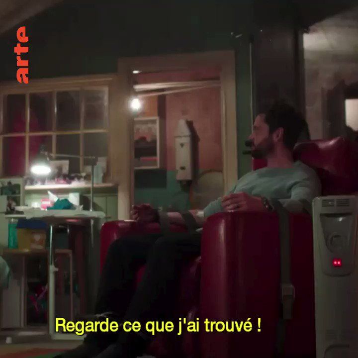 A screenshot showcasing arte subtitles. It is a man in an red armchair, within his living room.Top left is the logo for arte, in color red. The subtitle is yellow and reads "Regarde ce que j'ai trouvé", french for "look what I have found". The subtitles is at the bottom of the screen, over the armchair.