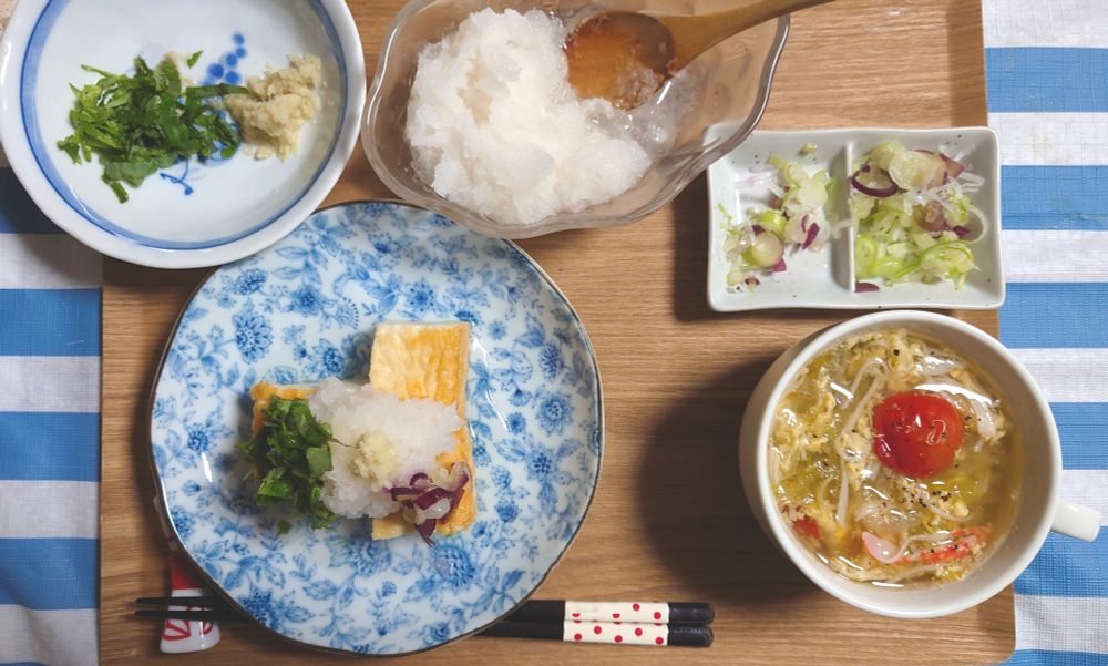 １人分の朝食が乗ったトレイ。大葉、。おろしショウガ、大根おろし、小口切りのネギと茗荷など薬味の皿があり、それらが焼いた絹揚げにたっぷりと乗っている。広めの白いカップには、千切りキャベツとカニカマ、ミニトマト、卵のコンソメスープ。