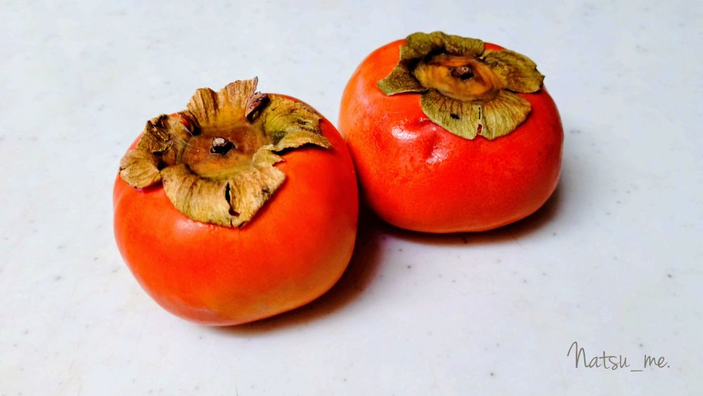 ご近所の庭に生った柿を頂いて、おやつに剥く前のまるまんまを撮影した写真。ヘタは大きく、実は小ぶりで少し傷や凹みがあるが、皮はつやつやとひかってとても鮮やかな橙色をしている。甘柿。