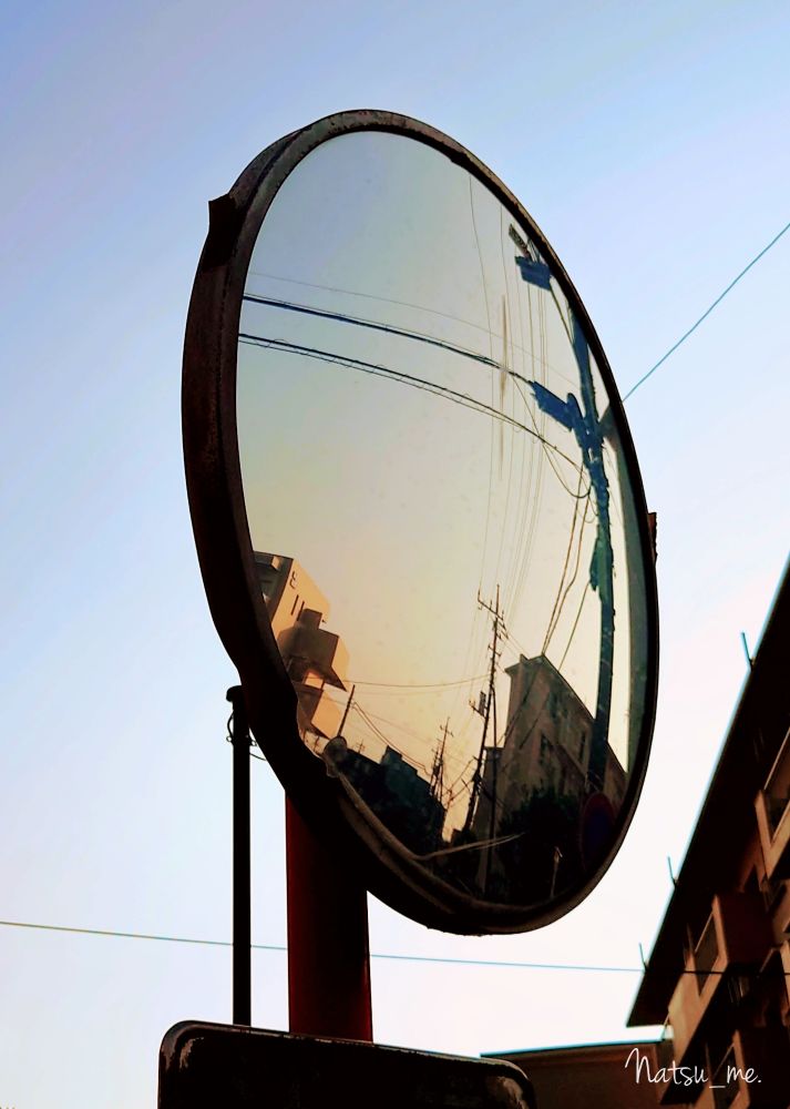 秋の夕方、暮れゆく空と夕陽に照らされる街並みがカーブミラーのなかに映り込んでいる写真。カーブミラーの外には薄く青が残る空、カーブミラーのなかには西の方角、夕陽が沈み太陽が街並みや電柱を照らすミニマムな世界が広がっている。