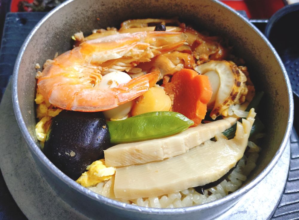 晩ごはんに宅配の釜飯セットを食べるところの写真。
蓋を取った釜の中には、炊きたての五目釜飯。
味付きごはんにボイルした殻付き海老、栗、飾り切りした人参、筍、椎茸、絹さや、うずら玉子、帆立などがきれいに盛りつけられている。