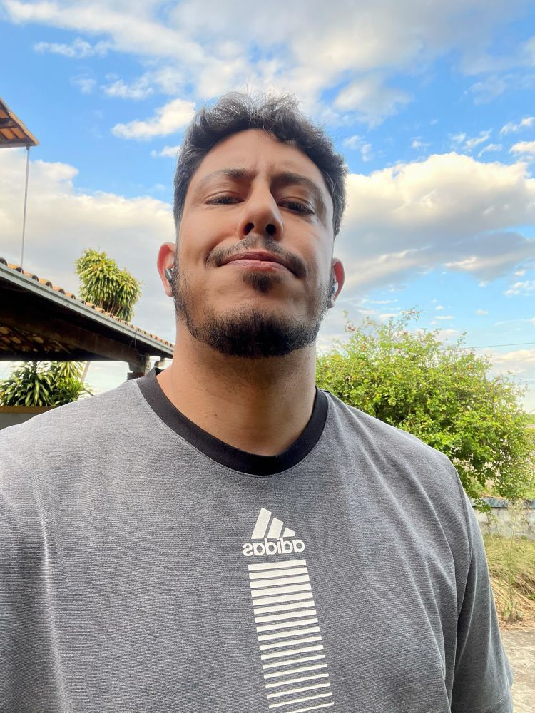 Auto retrato: eu, um homem moreno de cabelos e barba pretas usando uma camiseta cinza com detalhes em preto e branco escrito ‘adidas’, com um pé de acerola no fundo e o céu azul. 