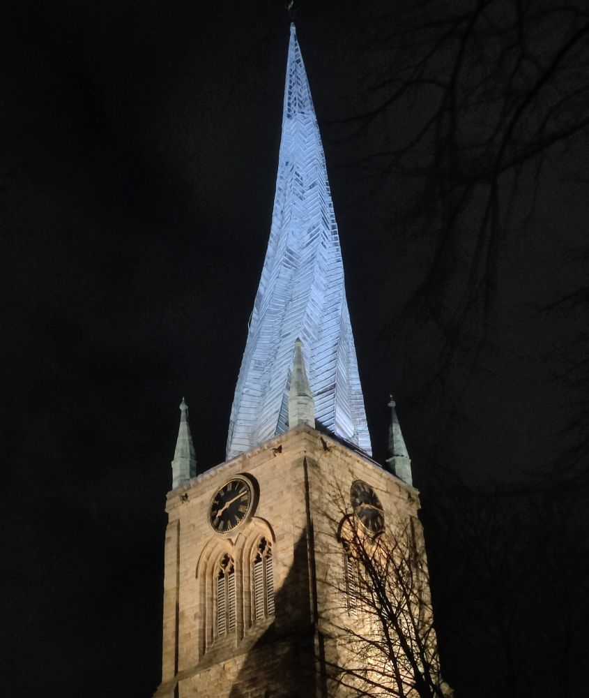 An English church spire lit in the darkness. The spire is. Ghostly pale grey blue. The spire is the famous Twisted Spire in Chesterfield, England. 
