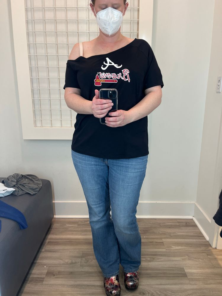 A photo of Ava, who is a white woman with purple hair and blue eyes. She is wearing blue jeans, an off the shoulder Atlanta Braves top with her right shoulder and a cream colored bra strap exposed, colorful Dansko clogs, and a white face mask. She is taking a selfie in the mirror of a white dressing room with faux wood flooring and excellent light. 