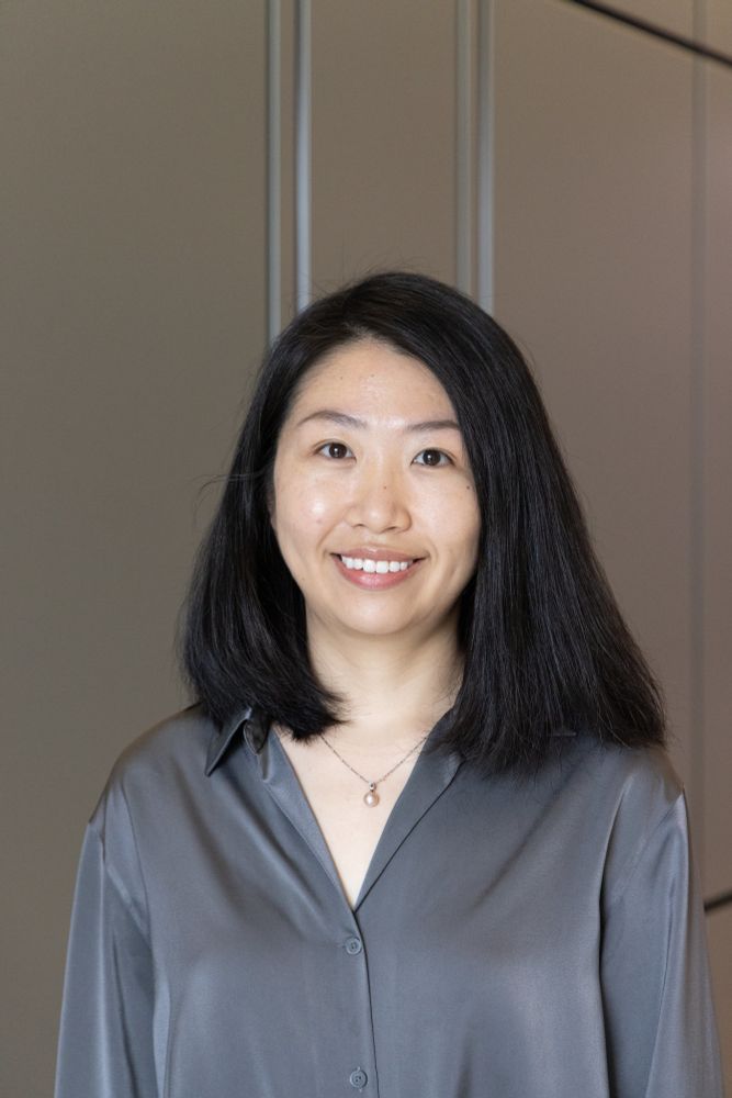 Xue stands against a gray background wearing a buttoned up blouse and hair down, smiling