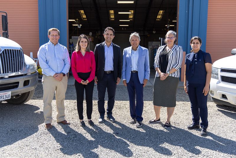 Six professionals stand in front of an industrial building entrance between two large white trucks on a sunny day.