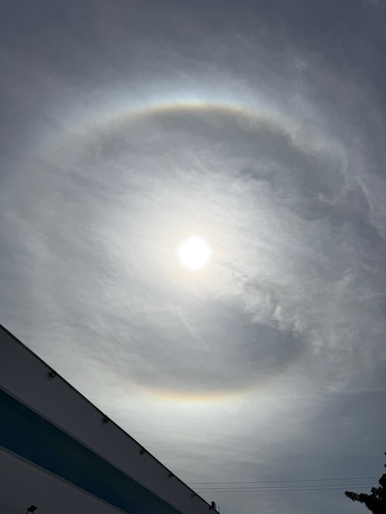 Foto do céu, com o sol ao centro e nuvens pouco espessas, causando um halo solar. O halo solar é uma espécie de arco-íris circular ao redor do sol, do ponto de vista de quem observa.