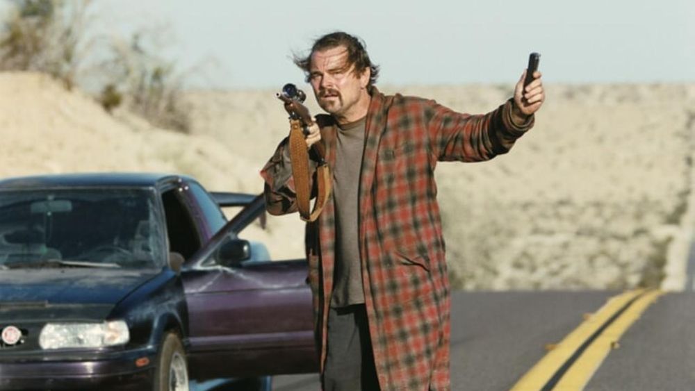 Leonardo DiCaprio als Pat „Ghetto Pat“ Calhoun / Bob Ferguson
mit wehenden Haaren, OliBa, kariertem, schäbigem Bademantel, T-Shirt hält einen Karabiner, zielt auf etwas und zeigt mit der anderen Hand ein (Erkennungs-)Gerät mit verzweifeltem Gesicht neben einem stehenden Auto auf einer Straße in der Wüste. 