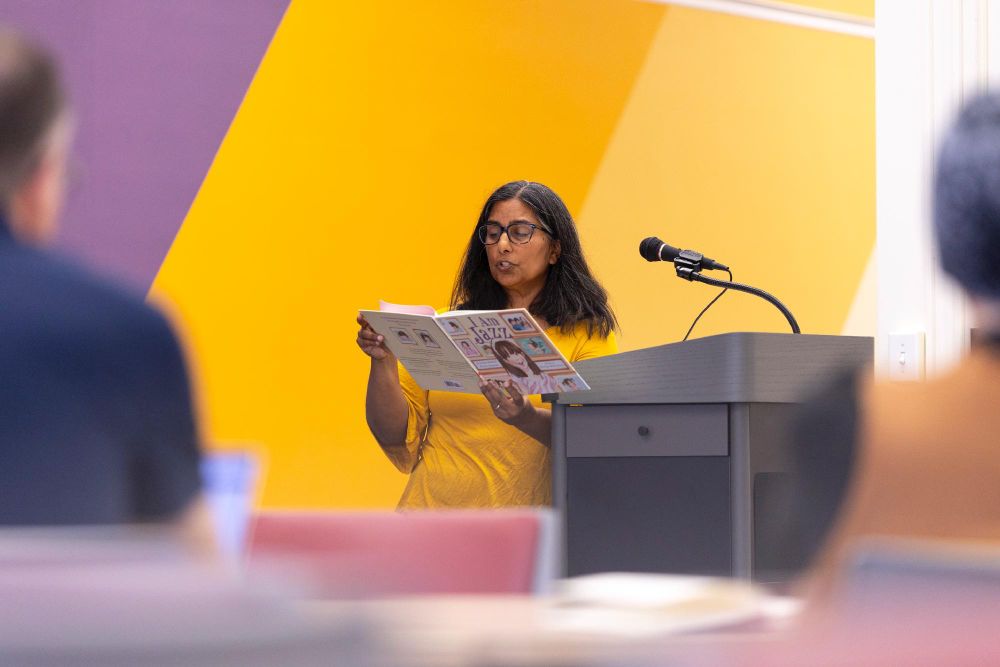 Person standing at a podium reading aloud from a newspaper in a room with yellow and purple walls.