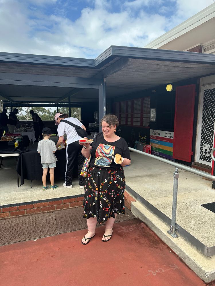 A woman stands holding a democracy sausage in one hand and a plate of cake in the other. She is wearing a democracy sausage shirt and a patterned skirt. In the background is a democracy sausage stall. 