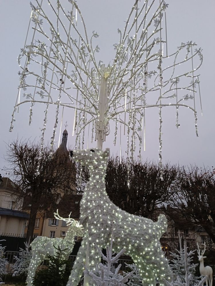 Un cerf blanc géant avec un arbre sur la tête à la place des bois. Le cerf et l'arbre sont couverts d'ampoules blanches.
Un autre cerf est visible en arrière plan.