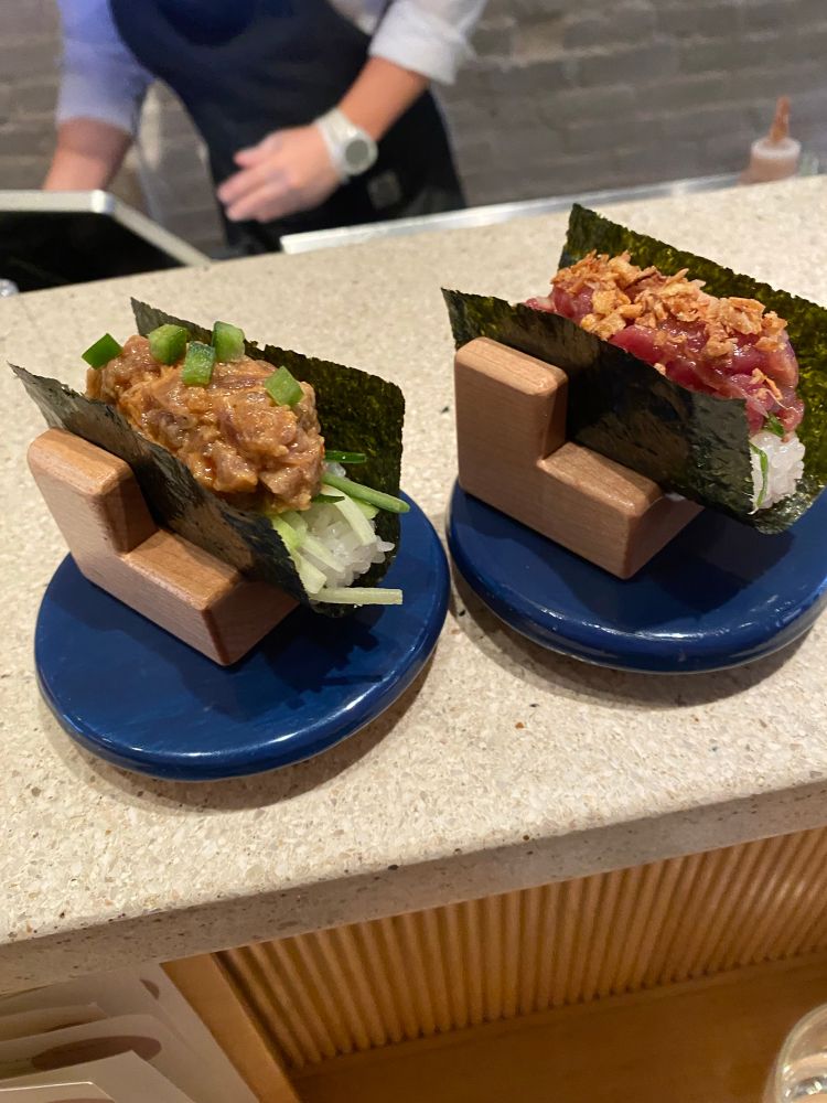 Two Japanese temaki rolls. One tuna poke style with crispy onion, and one spicy tuna with green chili pepper and cucumber. Both are served on sushi rice in a nori wrap.