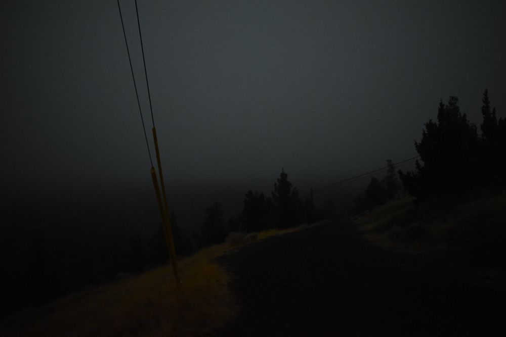 Photo of a gravel road leading down a hill at night in heavy fog. there is a few powerlines and dry grass leading down a steep drop-off into the fog to the side of the road