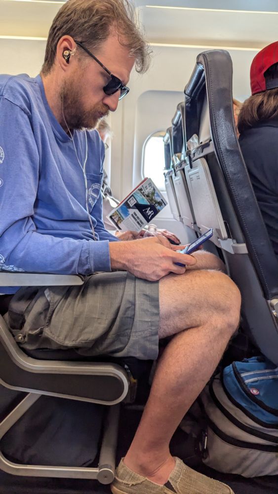 A tall man stuffed into an airplane seat, playing Balatro on a handheld gaming device.