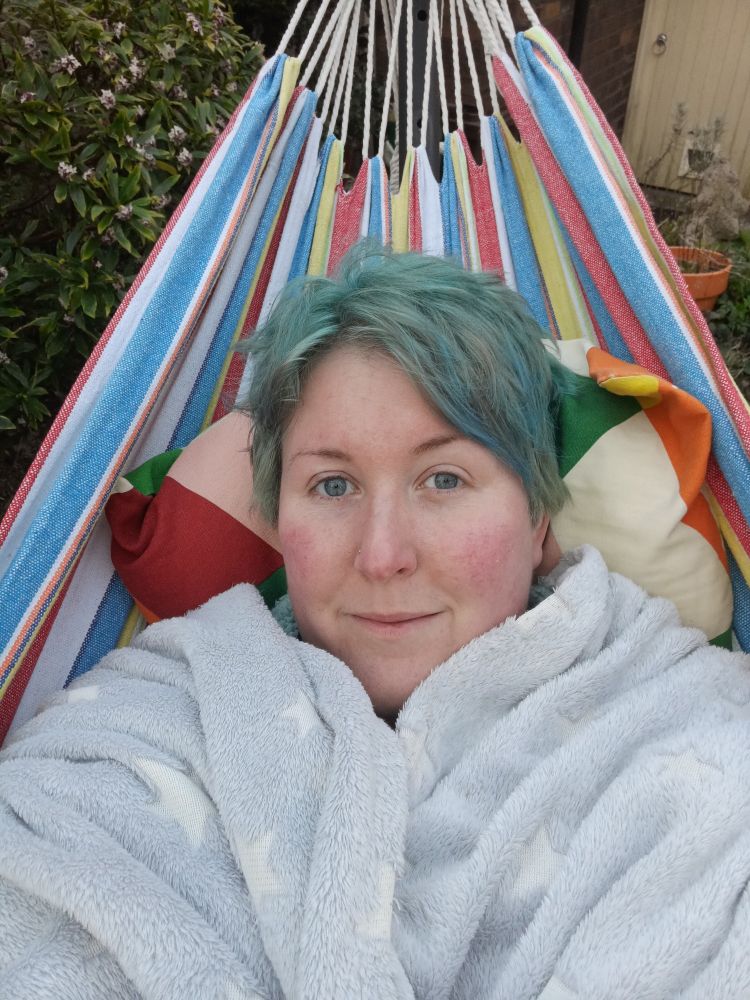 A white person with short seafoam blue hair and blue eyes smiles slightly at the camera. Only their head is visible, as they are wrapped in a pale grey and white starry blanket. Their head is propped up on a colourful cushion, and they are lying in a colourfully-striped hammock. Behind them, part of a back garden can be seen, including a shrub and a wooden door.