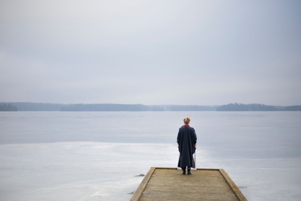 Pitkään takkiin pukeutunut hahmo seisoo puisella laiturilla. Jäinen järvi ja utuinen vastaranta.