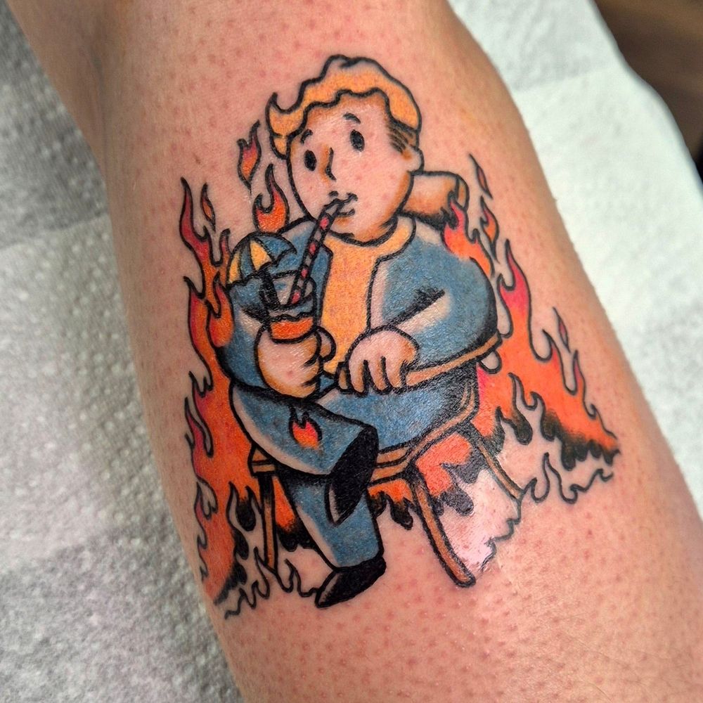A 3-inch tattoo on the forearm of Vault Boy, the mascot for the video game franchise Fallout, sitting on a chair sipping a cocktail whilst surrounded by flames.