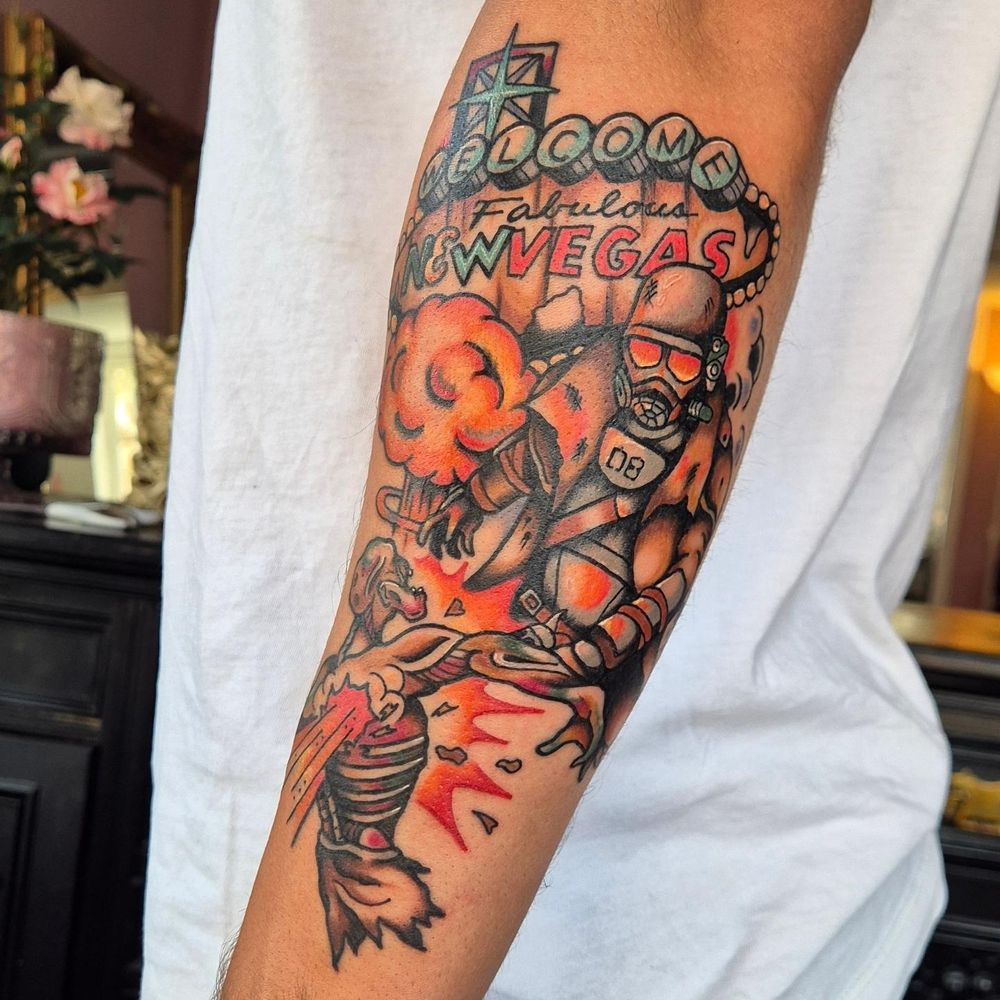 A large forearm tattoo based around imagery from the video game Fallout New Vegas. An NCR Ranger fires at a feral ghoul, with a mushroom cloud and the iconic New Vegas sign in the background. The tattoo is in American Traditional style, with bold lines and vivid colors.