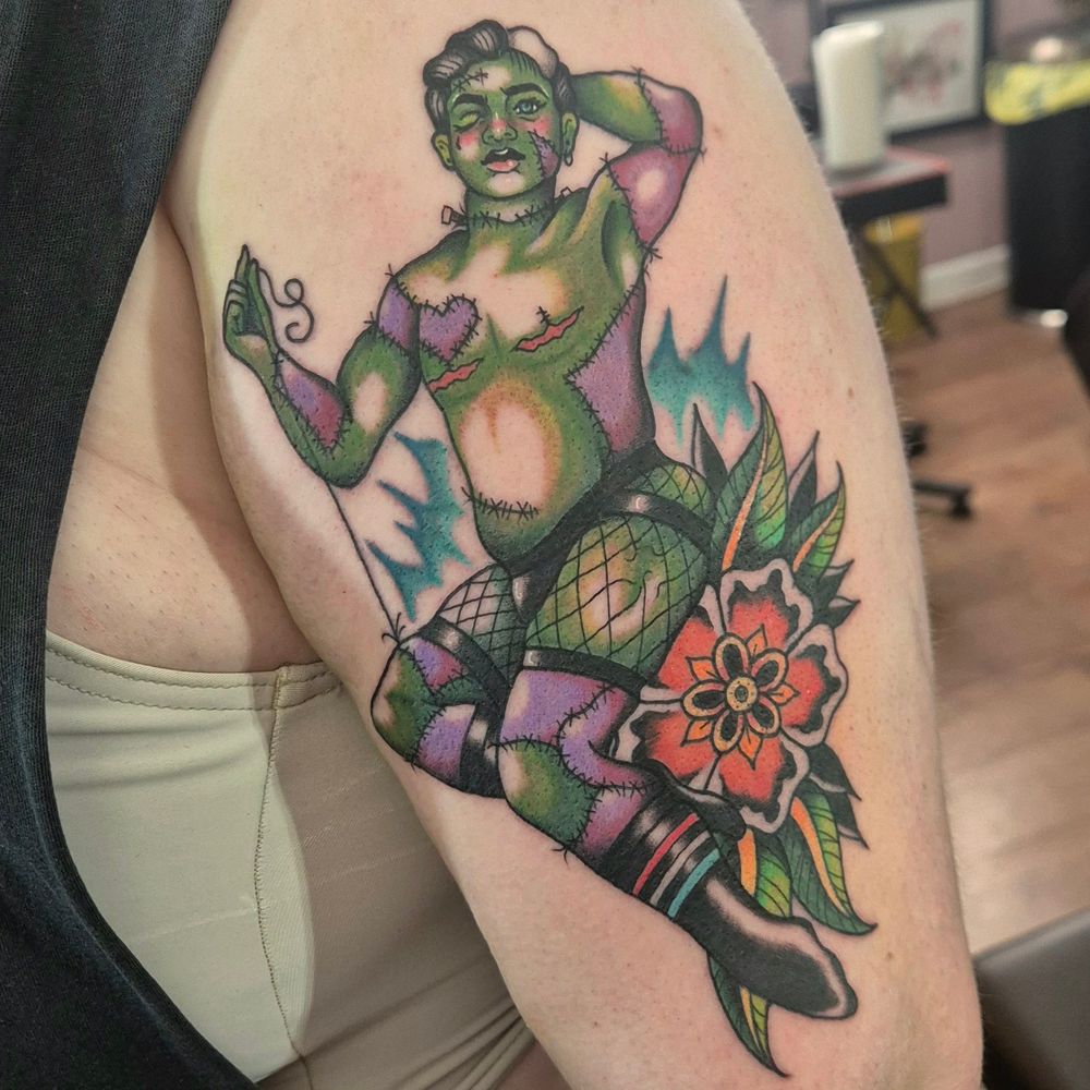 A large upper-arm tattoo of a transmasculine individual, in the style of Frankenstein's Monster. The colors are primarily greens, with a blue electric glow as a background, and a large orange flower. The pin-up wears a jock strap and ripped fishnet shorts, and is posed pulling a thread from his thigh. The tattoo is in American Traditional style, with bold colors and shading.
