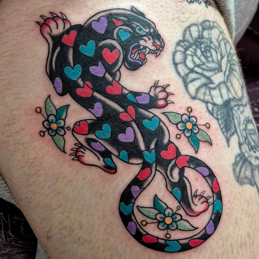 A large tattoo on the side of the thigh, of a crawling panther in American Traditional style. The panther has a pattern of hearts across it, in vivid pinks, blues and purples.