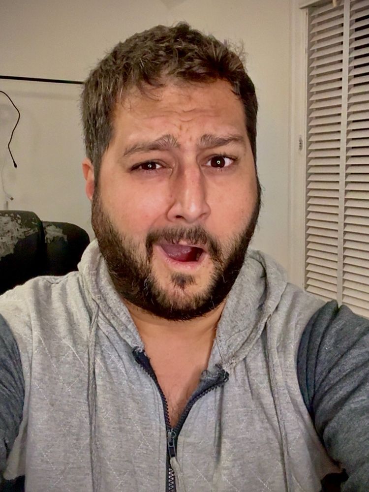 A middle aged, mixed race man wears a grey tracksuit onesie yawning into the camera with messy bed hair