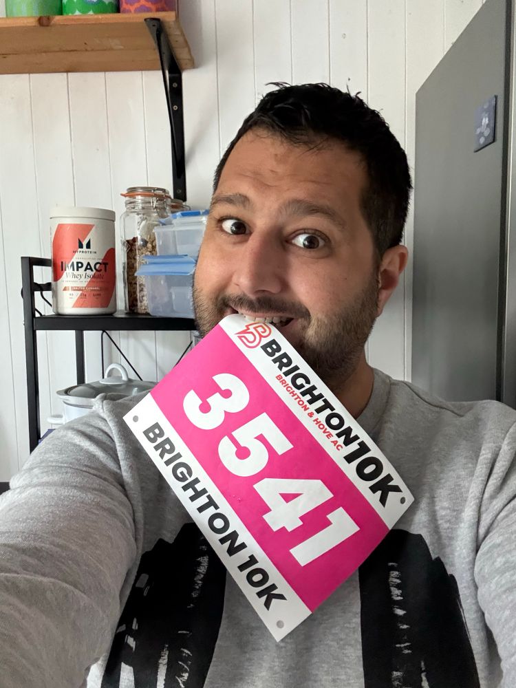 A brown, mixed race, middle aged man wearing a grey sweatshirt has a running bib in his teeth that says Brighton and Hove AC Brighton 10k: 3541
