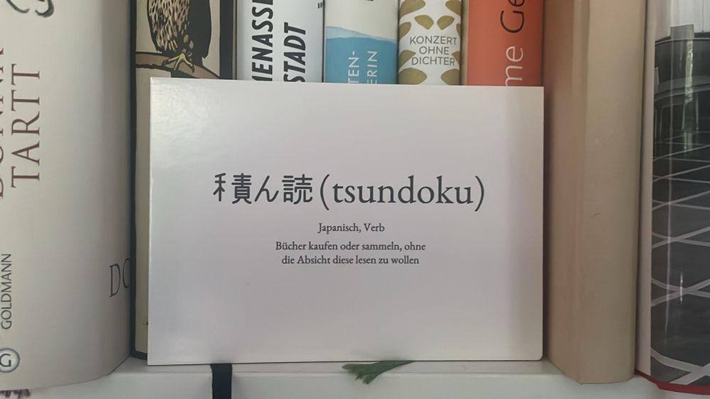 Postkarte, vor Büchern stehend; Aufschrift:

禾責誌（tsundoku）
Japanisch, Verb
Bücher kaufen oder sammeln, ohne die Absicht diese lesen zu wollen
