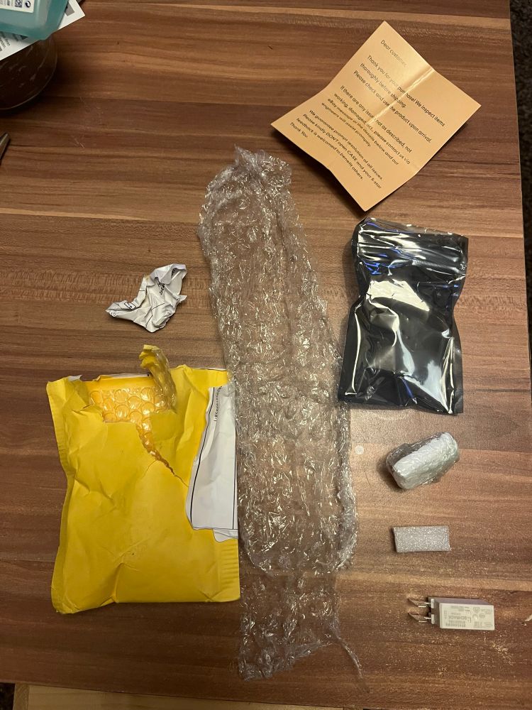 Lieferumfang eines Elektronikpakets auf einem Holztisch: geöffnete Versandtasche, Luftpolsterfolie, ESD-Schutzverpackung, kleiner in Folie verpackter Schaumstoffblock, loses elektronisches Bauteil,  Schaumstoffpolster und Hinweiszettel.