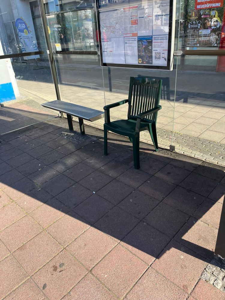 Bushaltestelle mit Glaswänden und Fahrplan an der Wand. Neben einer fest installierten Metallbank steht ein grüner Plastikgartenstuhl.