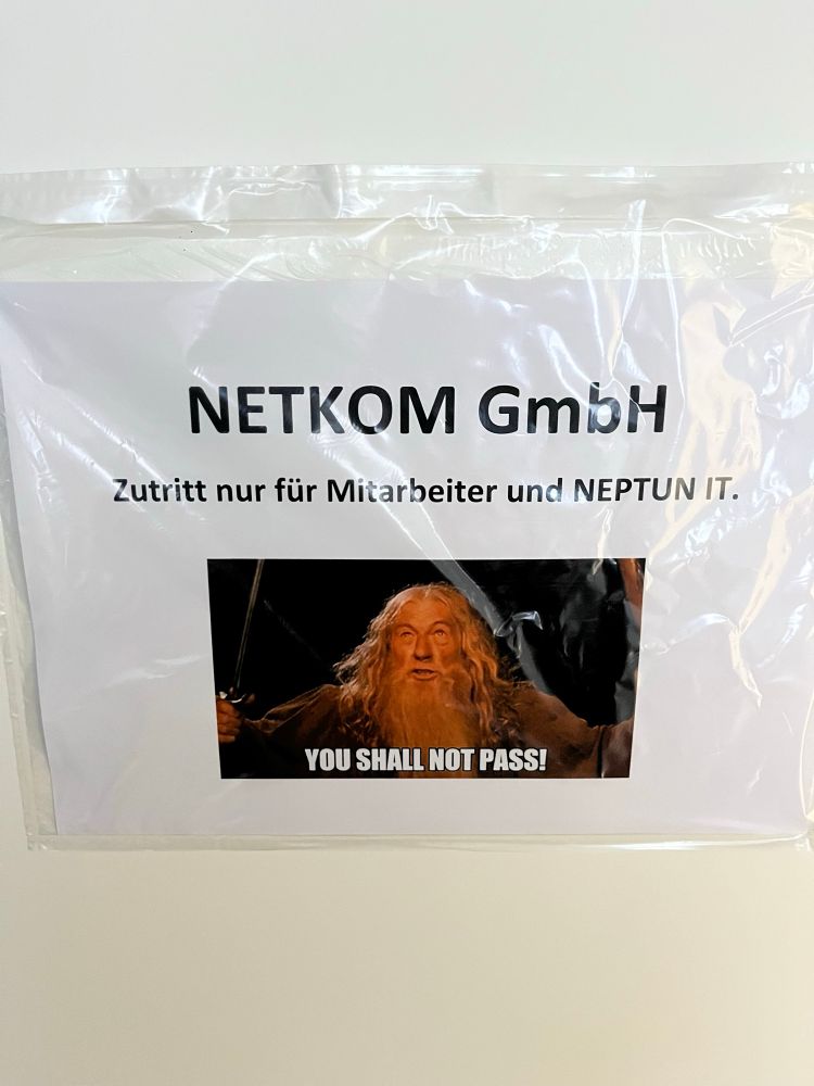 Ein ausgedrucktes Schild in einer Klarsichthülle hängt an einer hellen Wand. Oben steht: „NETKOM GmbH – Zutritt nur für Mitarbeiter und NEPTUN IT.“ Darunter befindet sich ein bekanntes Meme aus einer Fantasy-Filmszene: Ein älterer, bärtiger Mann in einem braunen Umhang hält einen Stab und ein Schwert erhoben und ruft energisch. Im unteren Teil des Memes steht der Text „YOU SHALL NOT PASS!“.