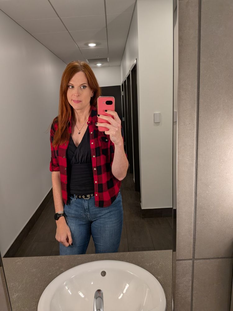 Nicole in a bathroom. She's wearing a red and black plaid shirt with a black wrap shirt underneath and jeans.