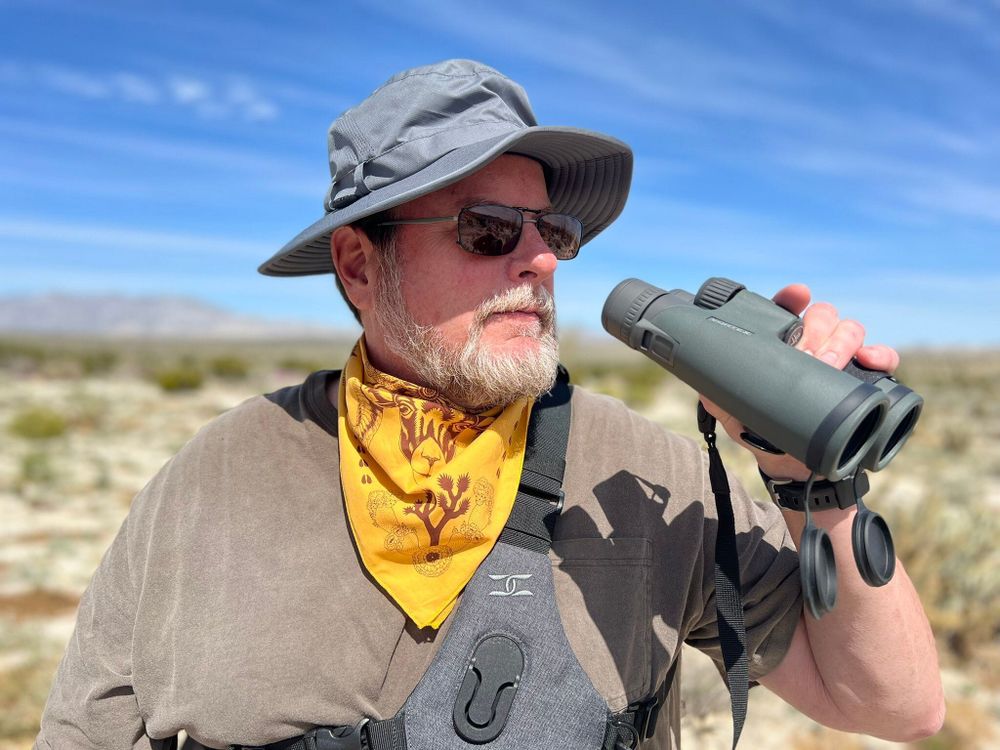 A handsome man (my husband, naturally) out in the desert birding holding binoculars, wearing sunglasses and a wide brimmed hat. He is wearing a desert animals. bandana around his neck in gold/yellow with brown ink showing a bighorn sheep and a joshua tree in this quadrant. 