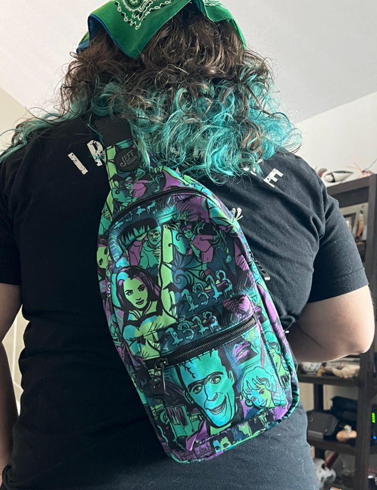 A small sling backpack with purple, black, teal and green color scheme with characters from the Munsters on it 