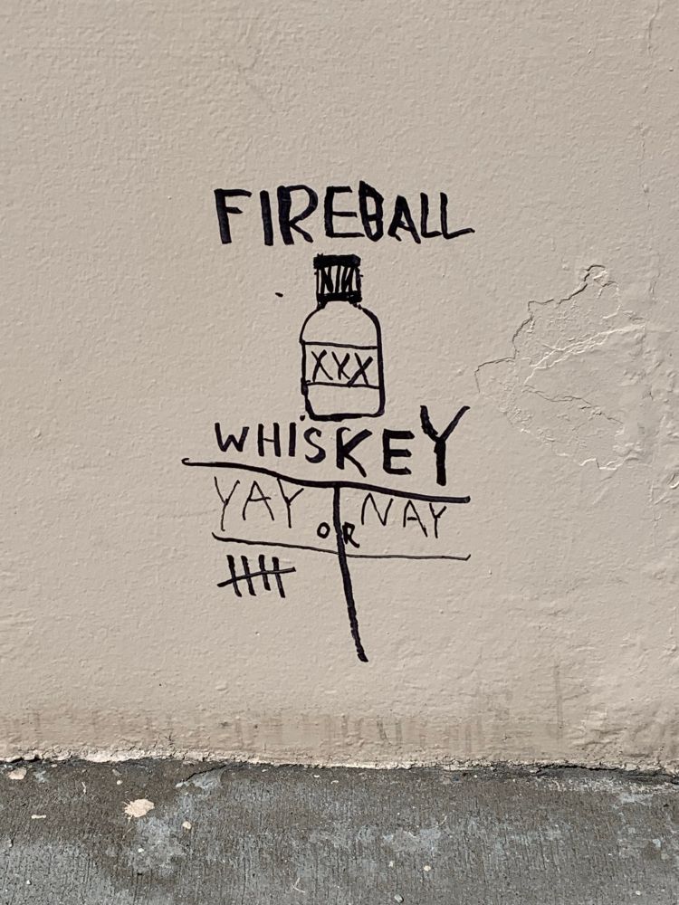 Fresh graffiti on a parking deck garage wall, inclusive of a stylized bottle of liquor, reads “Fireball Whiskey — Yay or Nay?” with five votes tallied in the affirmative and none in the other column.