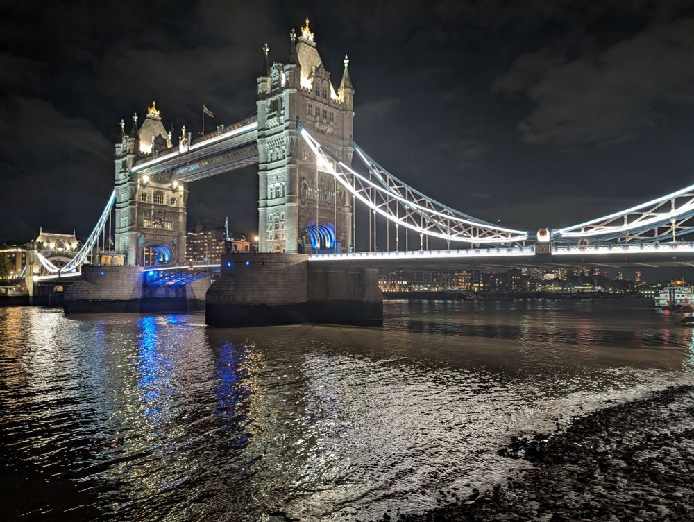 Tower Bridge illuminated at night. The lights reflect on the river and the foreshore. 