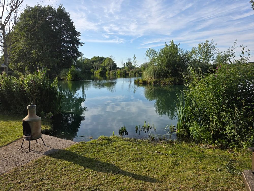 A photograph taken on a summer's day of a calm blue lake surrounded by trees and bushes. Grass and a chimenea line the side of the lake.