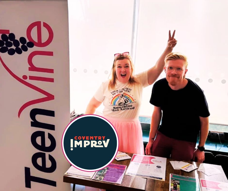 A photo of Grapevine's Sarah and Paul at the 'Full of Beans' event in May, standing next to a Teenvine banner and table with leaflets on it. They are both smiling. Sarah is wearing a unicorn rainbow t-shirt and pink skirt and making a peace sign above Paul's head. The Coventry Improv logo is also seen.