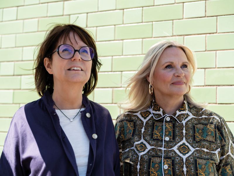 Two women stand side by side looking towards a clear sky outside the green tiled walls of Grapevine. The brunette woman wearing glasses is CEO Clare Wightman and the blonde woman wearing a patterned green dress is deputy CEO Mel Smith. Credit: Jorn Tomter for UCL Policy Lab magazine.