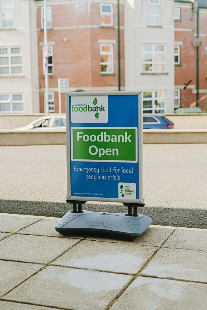 A blue and green 'Foodbank Open' A-board sign by the Trussell Trust stands on an urban pavement with cars on a road and a block of flats behind. Photo credit: Samuel Steele on Unsplash.