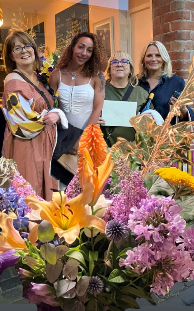Four women line up for a photo in a restaurant with a big bunch of flowers in the foreground. They are—left to right—Clare, Naomi, Cathy (holding a white envelope) and Mel.
