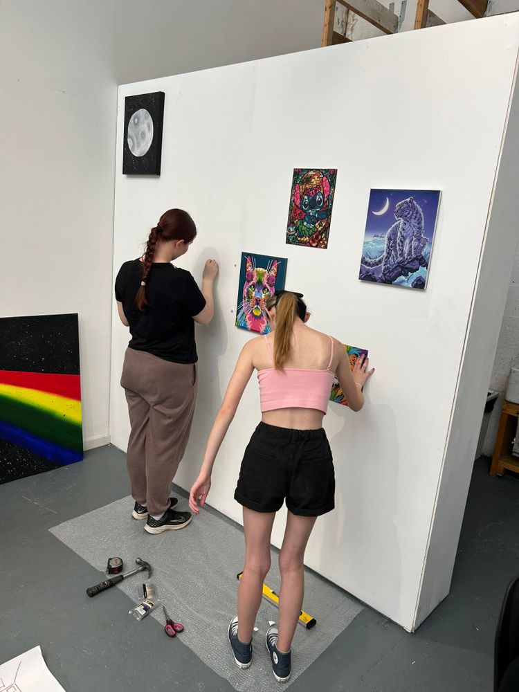 Two teenage girls use hammers and nails to put their art up on the white walls of the Art Riot Collective space.