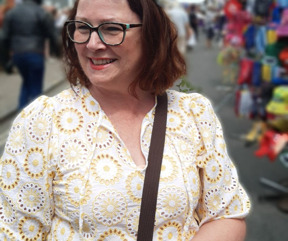 A white woman with brown shoulder-length hair and wearing glasses and a cream lace top smiles at a street event. She is Janet Doran.