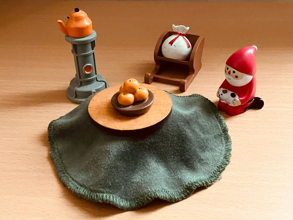 ミニチュアの冬っぽいセットが並べられている。

中央には小さなこたつ風の丸いちゃぶ台。その上には
みかんの盛り合わせが乗っている。

左奥には小さなやかんが乗ったミニストーブ。

右奥にはサンタさんのソリがあり、その上には袋が乗っている。

右側には赤い帽子のサンタさんがネコを抱いている。