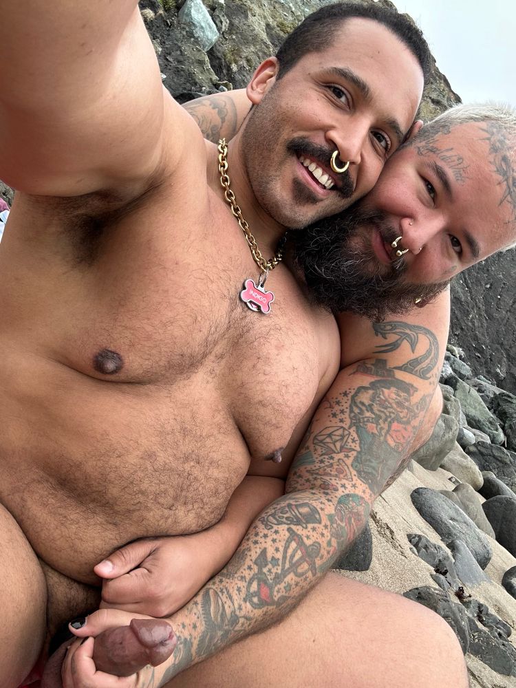 Two men naked at a beach, one is behind the other, grabbing the erect penis of the one in front