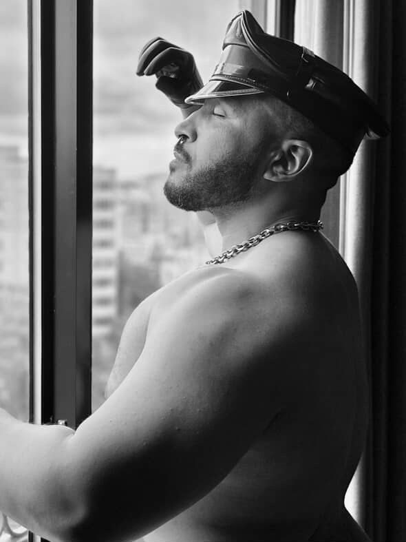 A black and white photo of a man wearing a leather cap and leather gloves, closing his eyes with his head posed a little bit elevated. He is standing at a window 