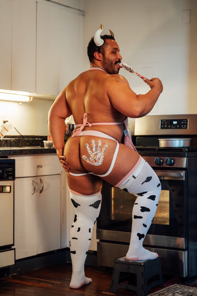 A professional photo shot of a big and muscular man in a kitchen setting, he has an icing hand print on his ass that has sprinkles on it, he is wearing thigh high cow print socks, bull horns, and a pink jockstrap and apron facing away from the camera, his head is turned back to see half of his face as he is licking a spatula 
