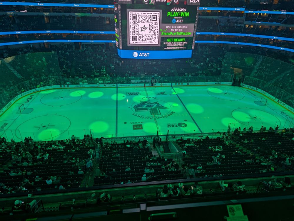 Picture of the American Airlines Center ice from the 300 terrace level on the night of the home opener. The crowd is beginning to fill in and there are green spotlights sweeping over the ice.