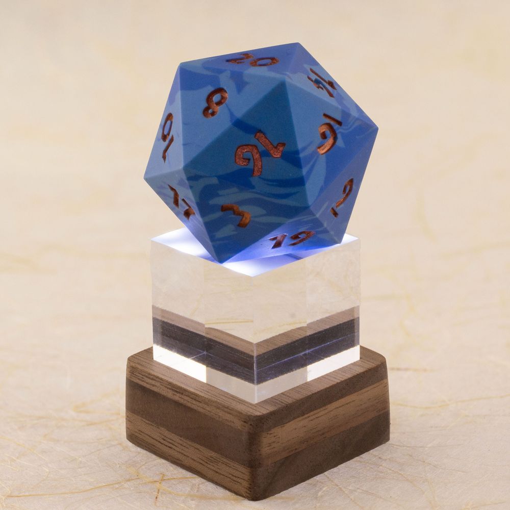 A one-inch clear acrylic cube sits in a small square layered walnut stand, which lights the cube and the 42 millimeter 20-sided die sitting on top of it from below. The die is a swirl of two shades of blue with metallic copper numbers.  Shown against a defocused background of handmade paper with fiber inclusions. Side view.